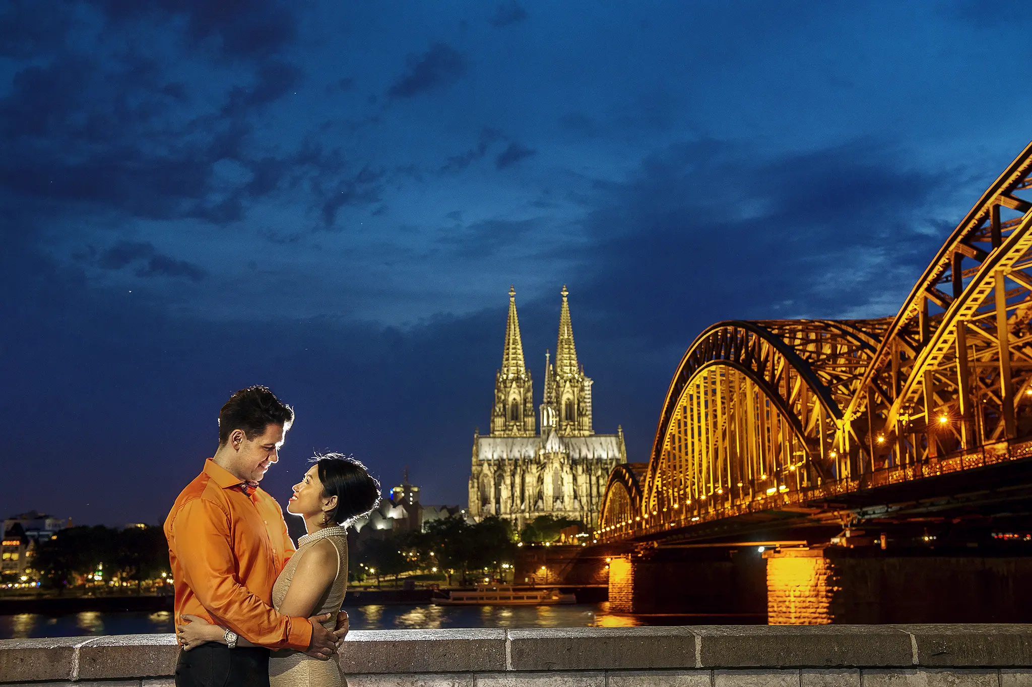 Cinematic pre-wedding portrait in Cologne, Germany, featuring a couple embracing at blue hour with the illuminated Cologne Cathedral (Kölner Dom) and Hohenzollern Bridge in the background